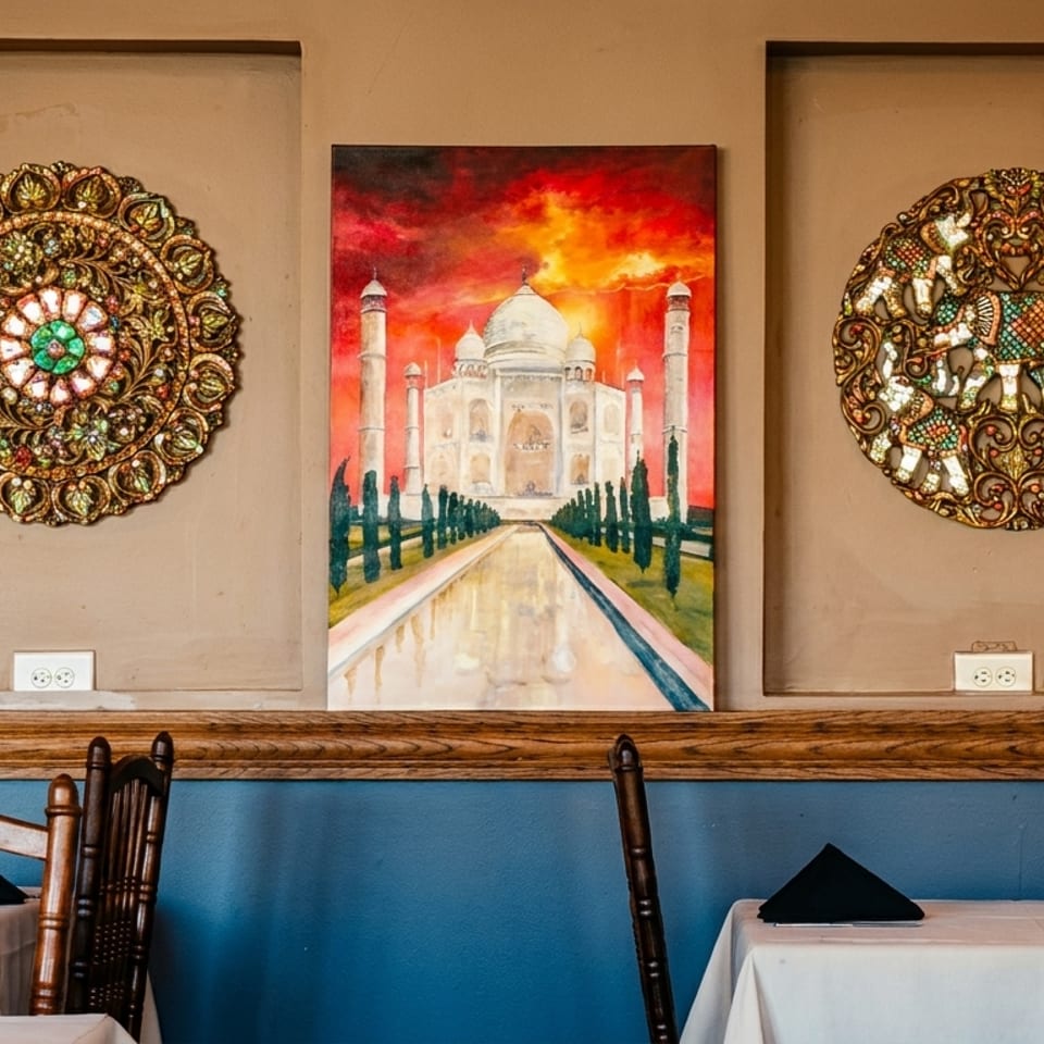 Warmly lit restaurant dining area with white tablecloths, dark napkins, ornate wall medallions, and a Taj Mahal painting.